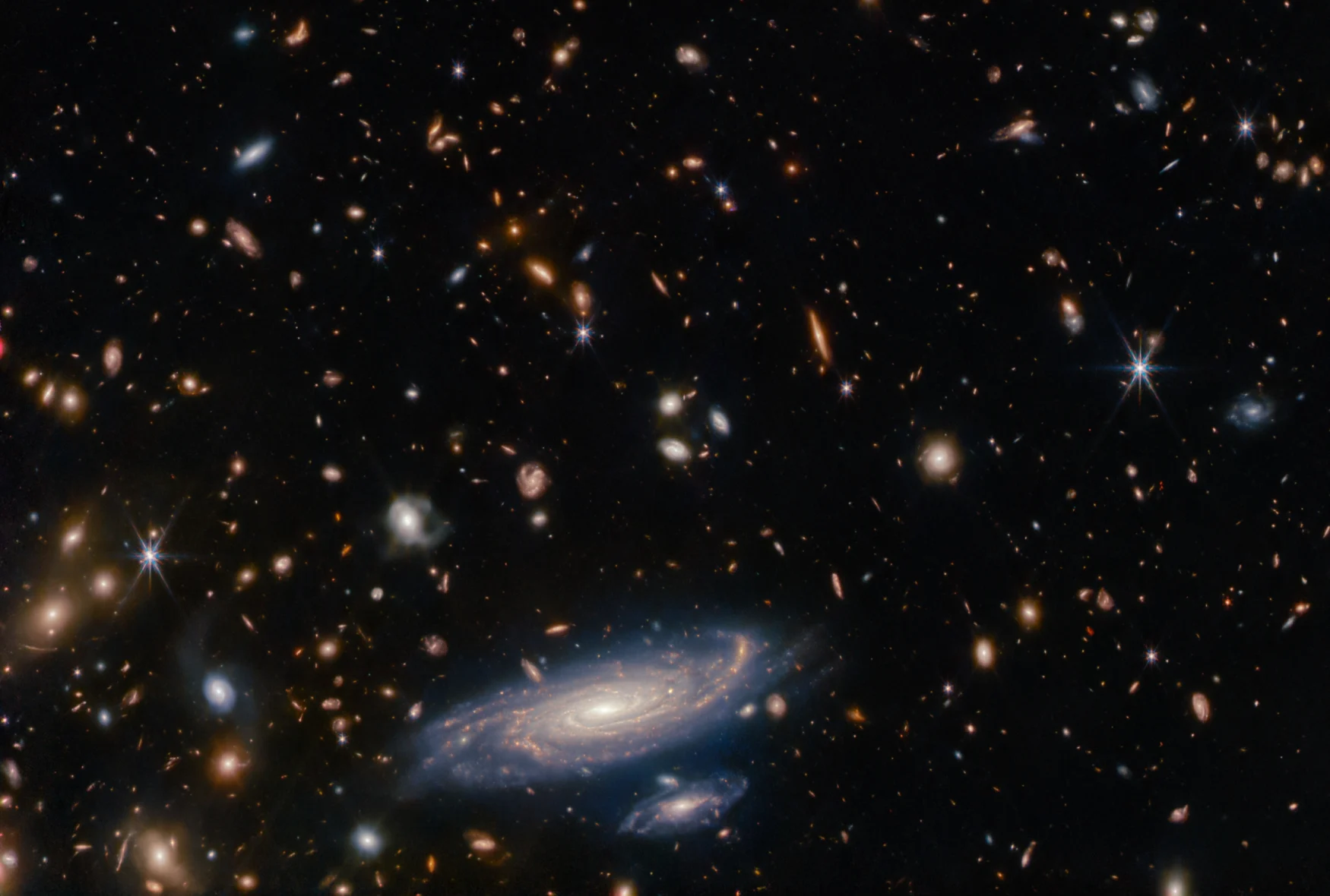 Image that was taken with the James Webb Space Telescope showing a spiral galaxy at the bottom center, surrounded by more distant galaxies (appearing as blobs) and individual stars.