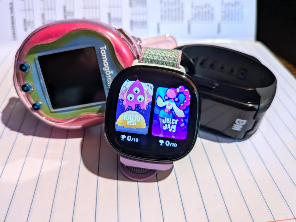 Fitbit Ace LTE pictured on an open composition notebook next to a pink Tamagotchi Uni and a black Digimon Vital Bracelet