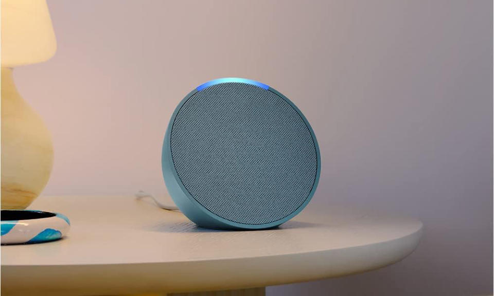 The Amazon Echo Pop speaker in blue sits on a night table with a yellow lamp nearby. 